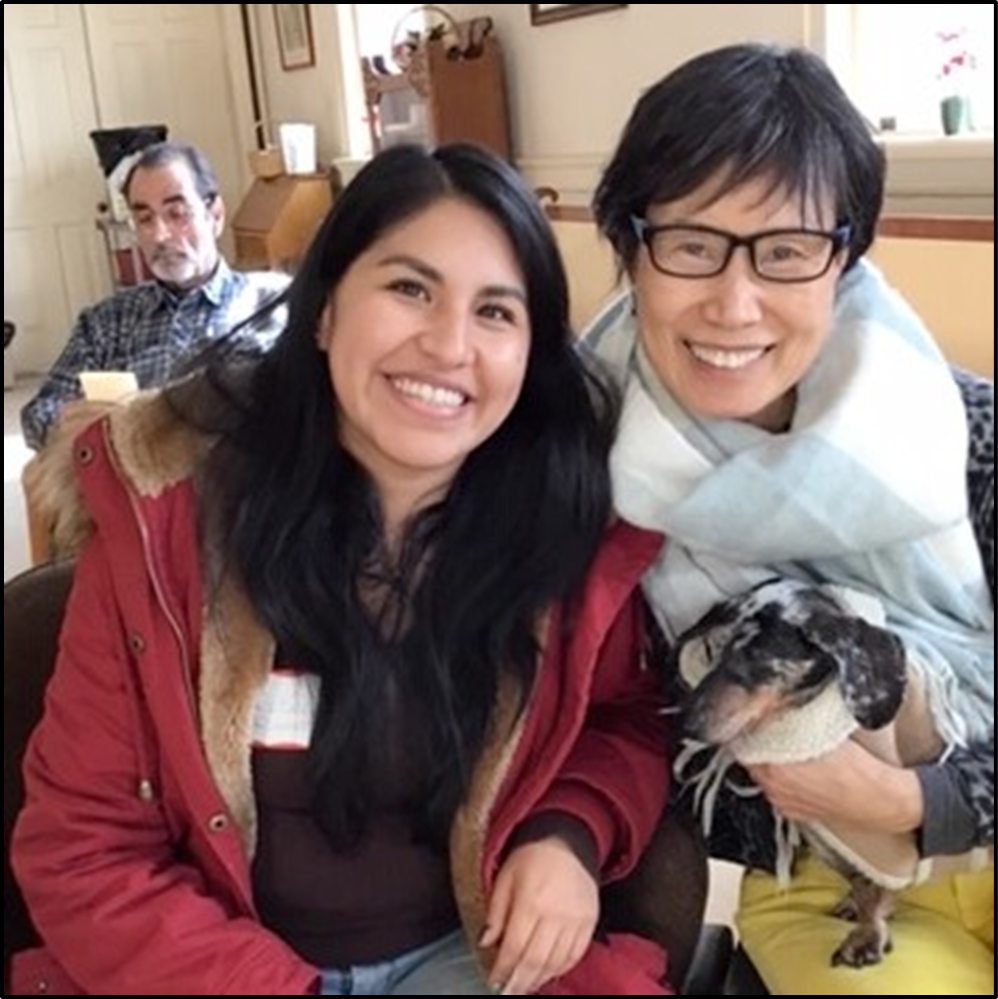 Two women smiling for the camera with a small dog, while a man sits in the background.