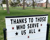 A sign expressing gratitude to service members, with the message "thanks to those who serve us all" displayed on a grassy background.