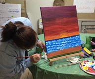 A person painting a colorful sunset over a body of water on a canvas.