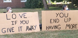 Two people behind large cardboard signs with the message "love... if you give it away, you end up having more" in a grassy area.