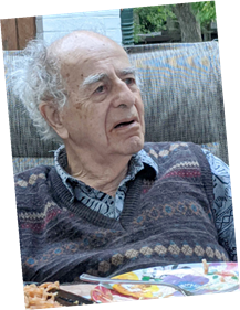 An elderly man with a thoughtful expression, wearing a patterned sweater, sitting outside on a couch.