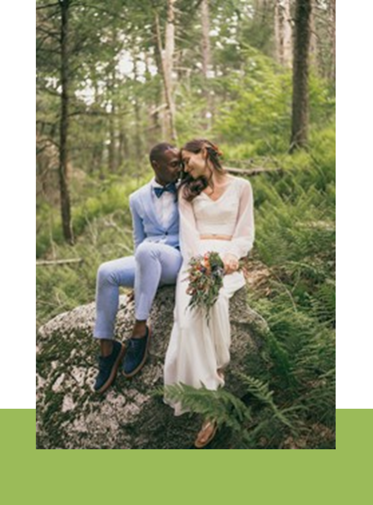 A bride and groom smiling at each other while sitting on a large rock in a lush forest. the bride holds a bouquet, and the groom wears a light blue suit.