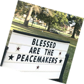 A sign with the message "blessed are the peacemakers" surrounded by stars, displayed in a park setting.