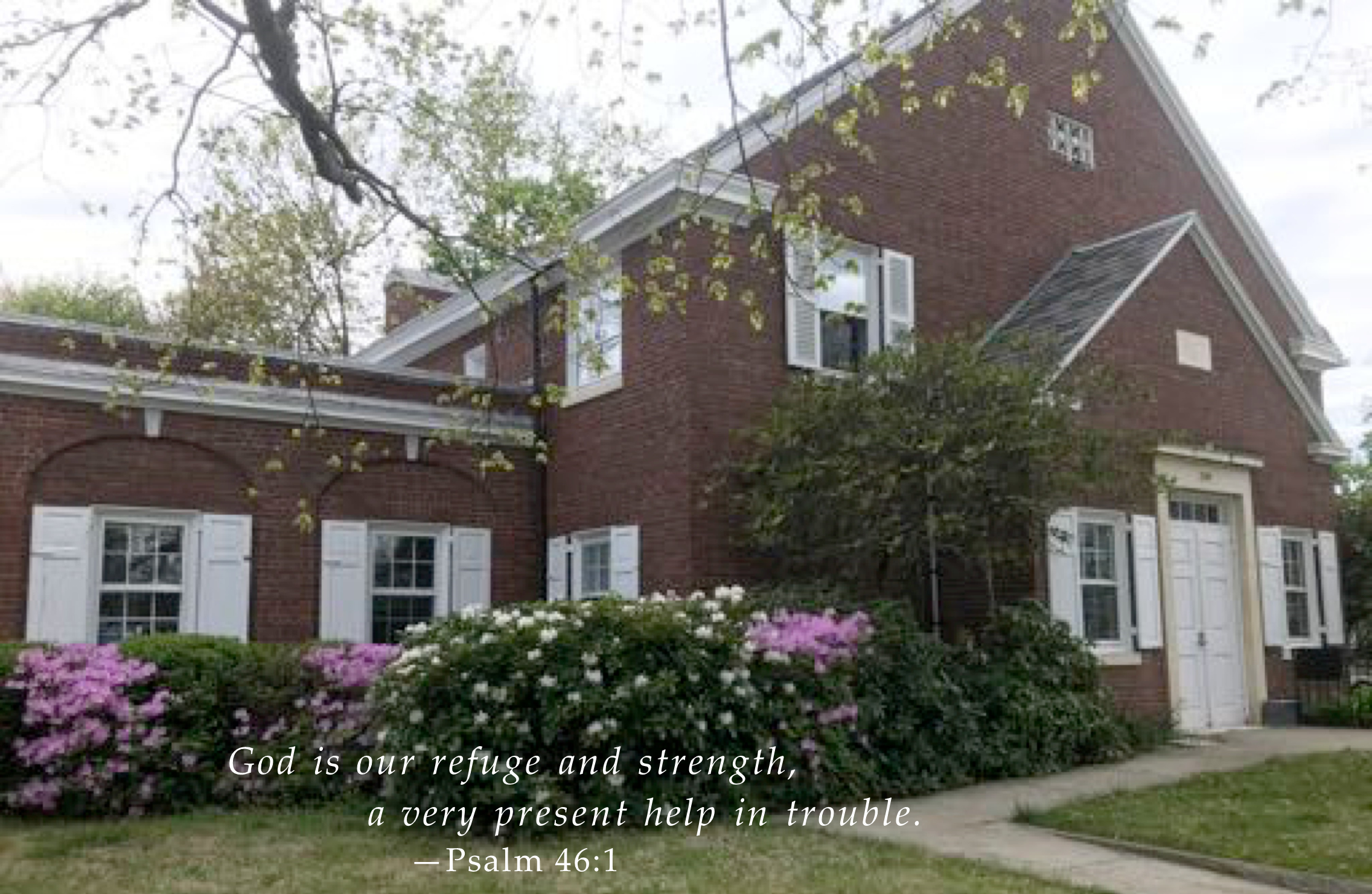 A quaint brick building with blooming bushes and a biblical verse, psalm 46:1, displayed on the front lawn.
