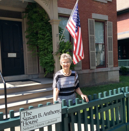 Woman standing in front of the susan b. anthony house, a national historic landmark, with an american flag displayed beside her.