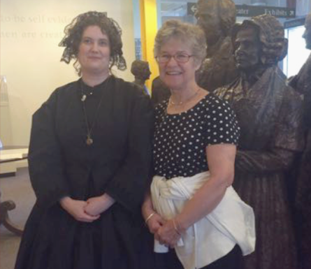 Two women, one dressed in historical costume and the other in modern clothes, smiling next to a bronze statue indoors.