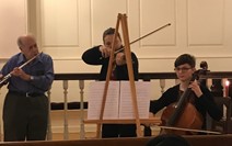 Three musicians performing with a flute, an easel with sheet music, and a cello in an indoor setting.